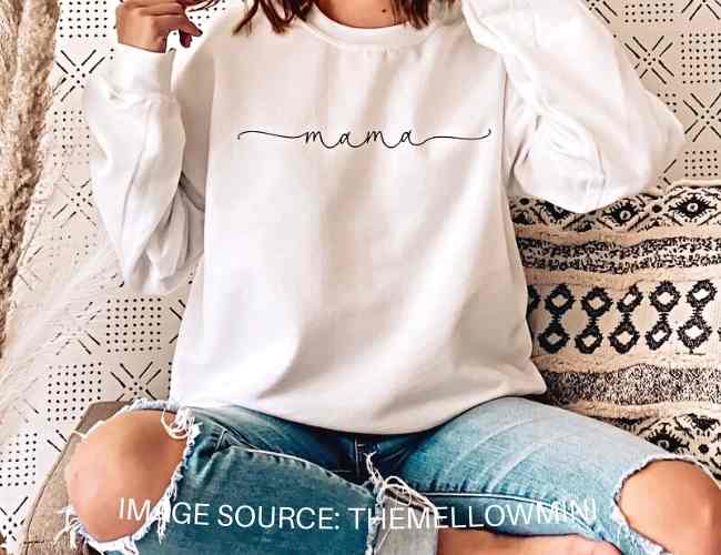 Woman sitting down wearing a white sweater that says mom, her head and feet are cropped off.